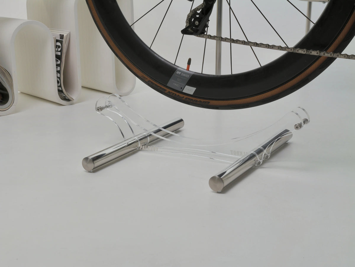 A clear acrylic and silver metal bicycle wheel stand supporting a bicycle wheel, set against a clean white background with a magazine in the blurred background.