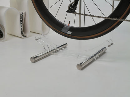 A clear acrylic and silver metal bicycle wheel stand supporting a bicycle wheel, set against a clean white background with a magazine in the blurred background.